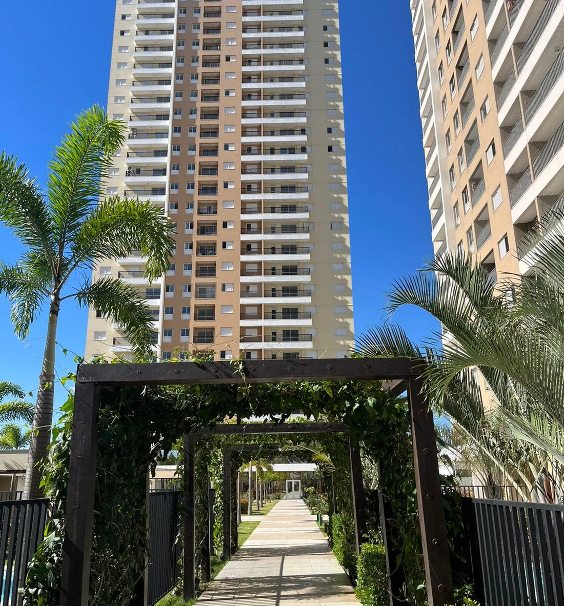 Torres de condomínio com palmeiras, pérgola e caminho ajardinado sob céu azul